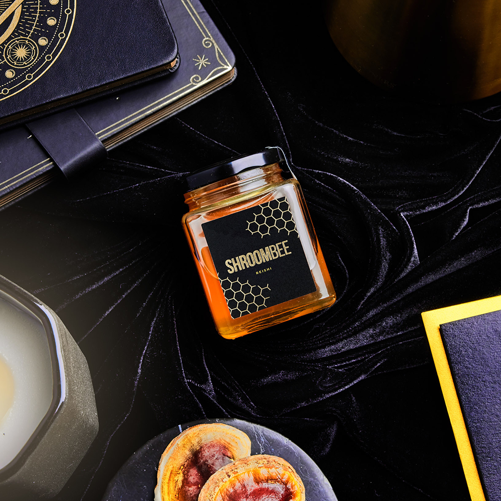 Jar of honey labeled 'Shroombee' on a dark surface with a notebook and food items.